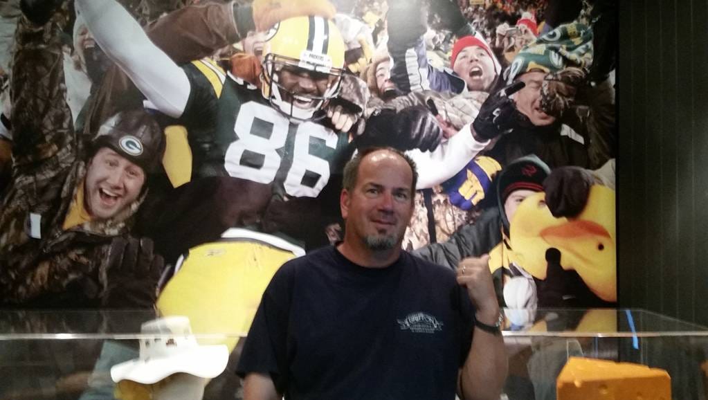 Doug Junk at the Pro Football Hall of Fame in Canton, Ohio standing in front of a photo of him, his son Augie and former Packer player Donald Lee during a Lambeau Leap in 2011.
